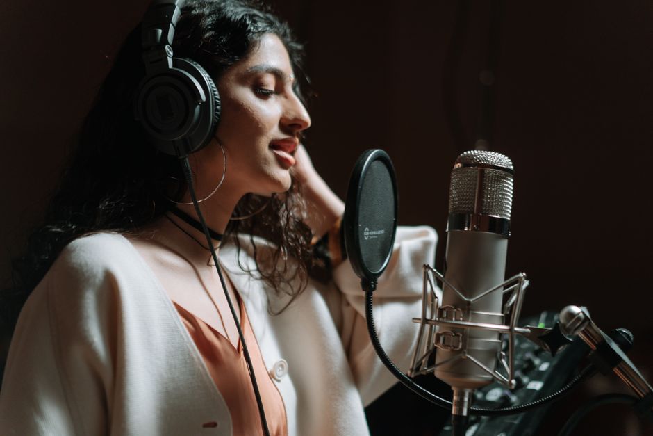 Woman in a music studio singing into a microphone, wearing headphones.